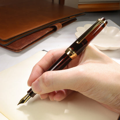 Hand holding a Mocha Brown fountain pen over a notebook with a brown leather wallet in the background.